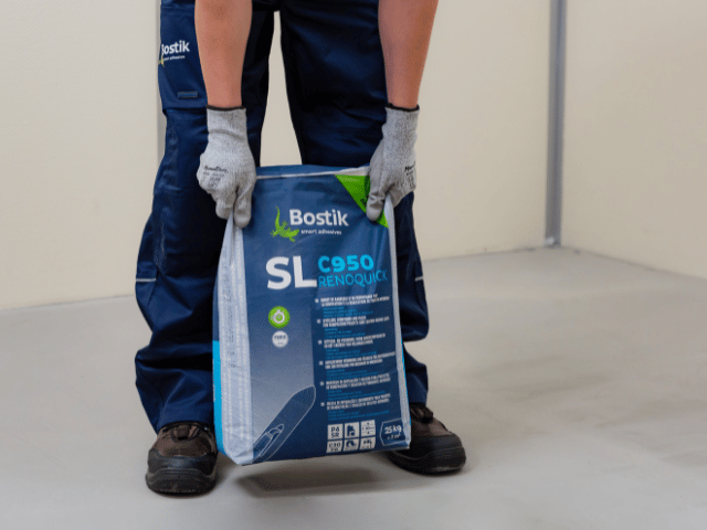 Worker holding a bag of Bostik SL C950 Renoquick floor levelling compound on a prepared concrete surface.