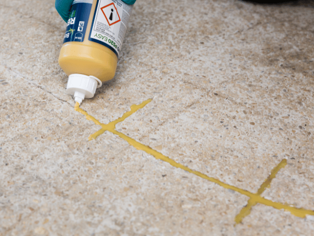 Bostik repair resin being applied from a squeeze bottle into marked cracks on a concrete floor, showing precise crack repair preparation.