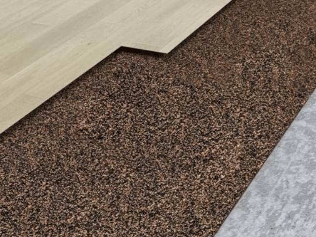 Layered flooring system showing a wood floor panel partially lifted to reveal a brown granulate sublayer installed over a concrete base using Bostik flooring solutions.