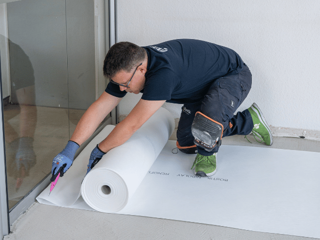 Bostik renovation membrane being unrolled and cut to size on a concrete floor near a glass door during subfloor preparation.