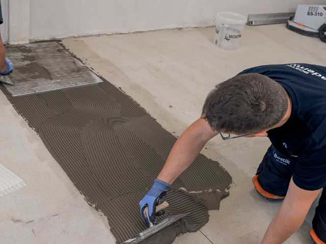 Bostik floor adhesive being spread with a notched trowel on a concrete subfloor during flooring installation process.