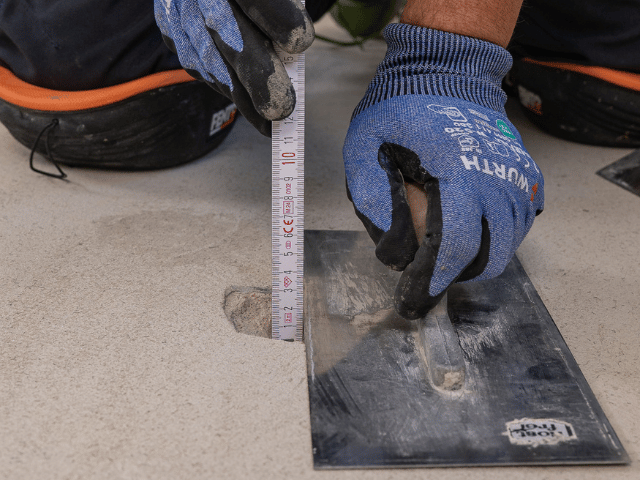 Worker measuring and filling a hole in a concrete floor using Bostik floor repair compound and a trowel.