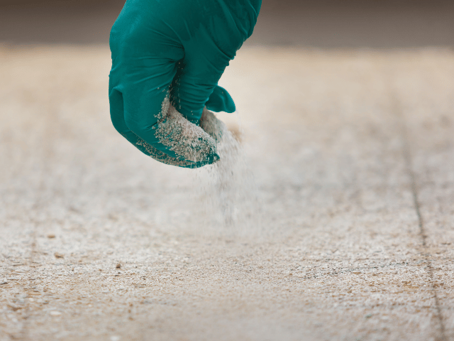 Bostik repair sand being sprinkled by hand onto a prepared concrete floor surface to improve adhesion before resin application.