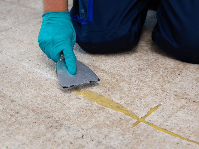 Bostik RENO repair material being smoothed with a scraper over filled cracks in a concrete floor to create a level surface.