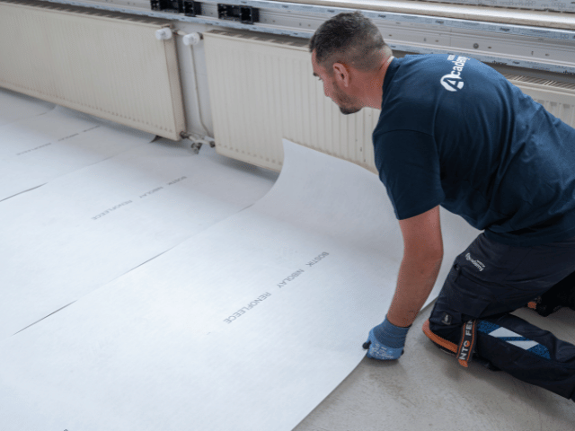 Bostik renovation membrane being positioned and rolled out across a prepared subfloor for flooring installation.