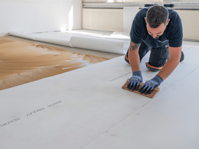 Bostik renovation membrane being pressed and smoothed onto adhesive using a hand tool to ensure proper bonding on a concrete subfloor.