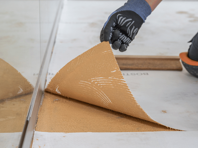 Close-up of Bostik floor adhesive being lifted from a subfloor surface, showing adhesive coverage and bonding layer during installation.