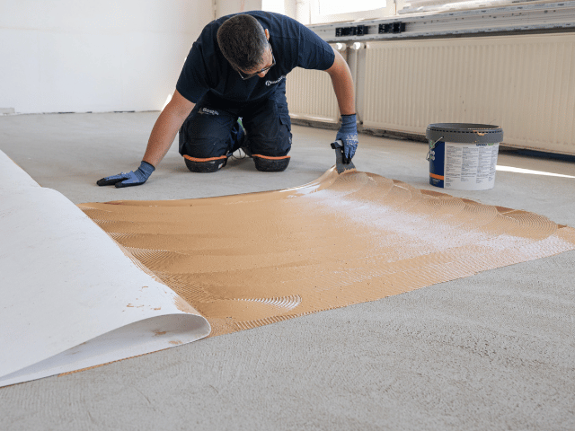 Bostik floor adhesive being evenly spread with a trowel across a concrete subfloor, with a bucket of adhesive nearby during flooring installation preparation.
