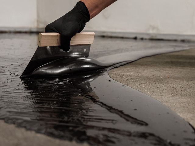 Bostik waterproofing compound being spread with a trowel across a concrete floor surface to create a protective coating.