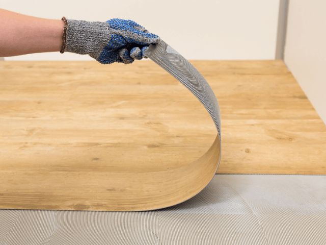 Flexible flooring panel being lifted to reveal adhesive application underneath, demonstrating installation with Bostik flooring systems.